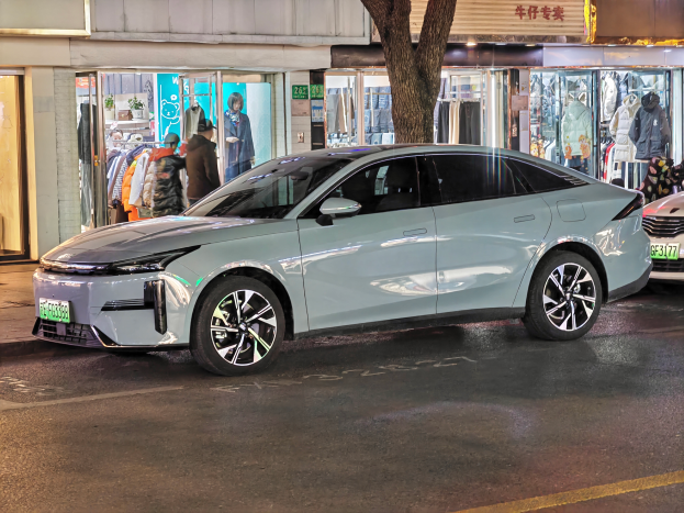 Ein Mitsubishi Lancer EV-Elektroauto, das nachts auf einer Straße geparkt ist, umgeben von Gebäuden, Geschäften, Schaufensterpuppen mit Kleidern, beleuchteten Schildern und anderen städtischen Elementen.