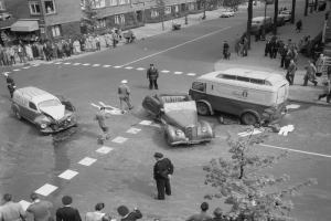 Schwarz-weiß-Szene eines Autounfalls an einer Straßenkreuzung mit mehreren Fahrzeugen, darunter ein Lieferwagen, und einer Gruppe von Zuschauern, mit Gebäuden, Bäumen, Strommasten und Texttafeln im Hintergrund.