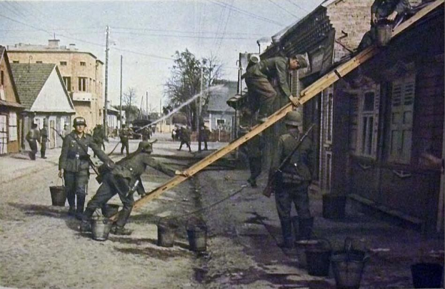 Deutsche Soldaten im Zweiten Weltkrieg in Deutschland stehen auf einer Straße neben einem Gebäude, tragen Helme und halten hölzerne Gegenstände, mit Eimern auf dem Boden und Gebäuden, Bäumen, Polen, Drähten im Hintergrund unter einem klaren Himmel.