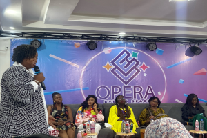 Eine Frau, die Gründerin des Opernveranstaltungsorts, steht mit einem Mikrofon vor einer Gruppe sitzender Frauen, mit Tischen voller Flaschen und anderen Gegenständen im Vordergrund und einem Banner mit Text und Deckenbeleuchtung im Hintergrund.