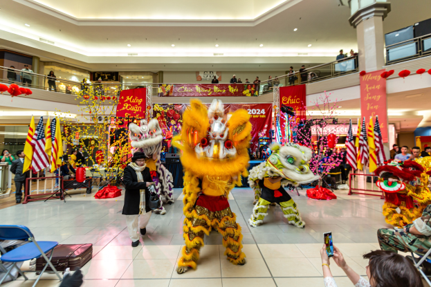 Ein farbenfrohes chinesisches Neujahrsfest in einem Einkaufszentrum mit Menschen in Kostümen, die tanzen, anderen, die auf Stühlen sitzen und Handys halten, mit Fahnen, Bannern, Dekorationen, Geländern, Säulen, Deckenlampen und einem Wasserzeichen unten.