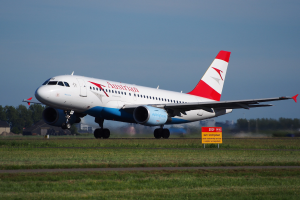 Airbus A320-200 der Austrian Airlines startet von Frankfurt Airport, mit einem Schild im Vordergrund, Gras darunter, Bäumen und Gebäuden im Hintergrund und einem klaren blauen Himmel.