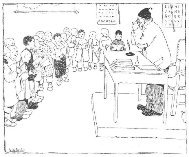 Ein Mann sitzt an einem Schreibtisch mit Büchern und Gegenständen vor einer Gruppe von Kindern, mit Papieren an der Wand dahinter, trägt Schuhe.