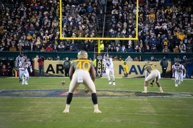 Fußballspiel in einem Stadion mit Spielern in Sportkleidung und Helmen im Vordergrund und Zuschauern im Hintergrund, einschließlich Pfosten und Tafeln mit Text.