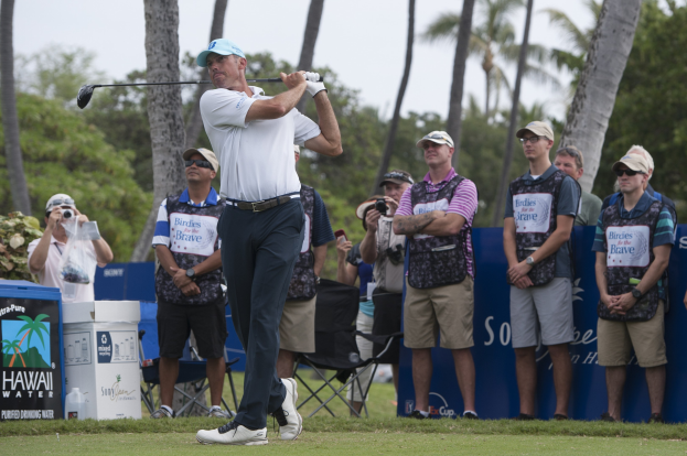 Ein Mann in einem weißen T-Shirt, schwarzen Hosen und einer blauen Mütze schwingt einen Golfschläger auf einem saftig grünen Feld, mit Zuschauern und Kameras dahinter, vor einem Hintergrund aus Bäumen und einem klaren blauen Himmel.