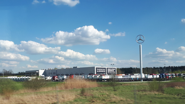 Eine Mercedes-Benz-Fabrik in Deutschland mit Fahrzeugen auf der Straße, umgeben von Grünflächen und Gebäuden unter einem bewölkten Himmel.