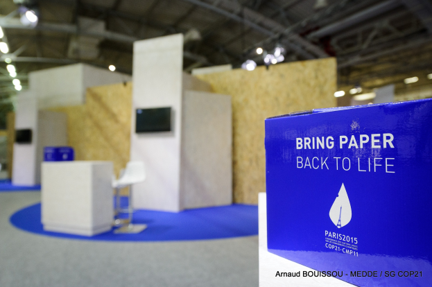 Ein Messestand mit einem blauen und weißen Schild, auf dem 'Bring Paper Back to Life' steht, auf der rechten Seite, im Hintergrund leicht unscharfe Stühle, Tische, ein Wandfernseher und Deckenleuchten.