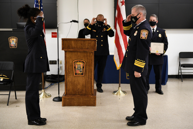 Gruppe uniformierter Personen bei einer Vereidigung eines neuen Feuerchefs, die ein Zertifikat halten, mit Fahnen, Podium und maskierten Teilnehmern im Hintergrund.