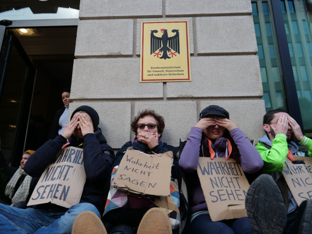 Eine Gruppe von Menschen sitzt vor einem Gebäude und hält Schilder, einige decken ihre Gesichter ab, mit einem Logo und einer Tafel an der Wand und einer Glastür auf der rechten Seite.