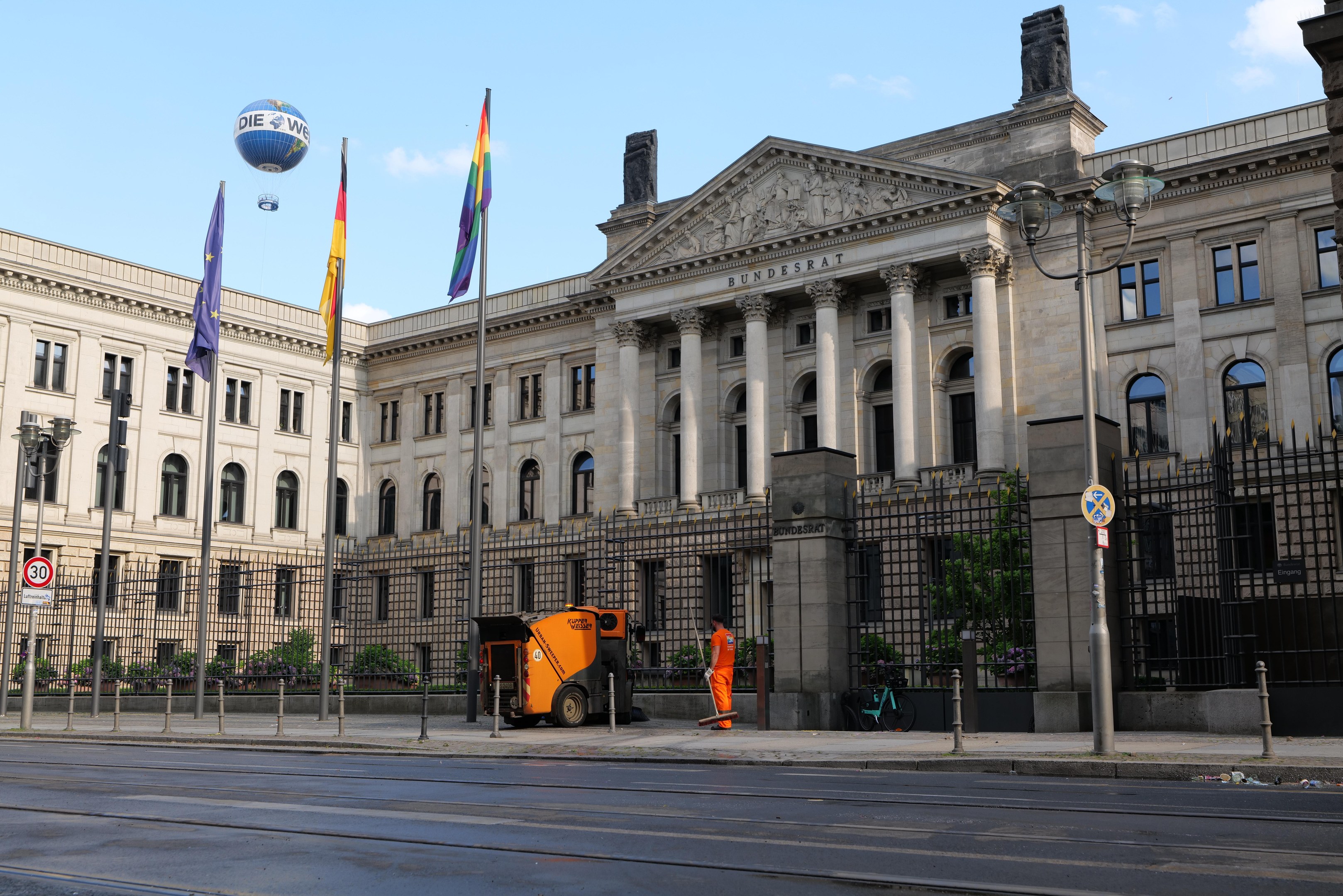 Großes modernes Regierungsgebäude (Bundestag) in Berlin mit vielen Fenstern, umgeben von Straßeninfrastruktur, Fußgängern, Fahnen und Bäumen unter einem bewölkten Himmel.
