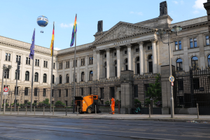 Großes modernes Regierungsgebäude (Bundestag) in Berlin mit vielen Fenstern, umgeben von Straßeninfrastruktur, Fußgängern, Fahnen und Bäumen unter einem bewölkten Himmel.