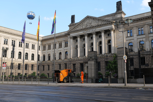 Großes modernes Regierungsgebäude (Bundestag) in Berlin mit vielen Fenstern, umgeben von Straßeninfrastruktur, Fußgängern, Fahnen und Bäumen unter einem bewölkten Himmel.