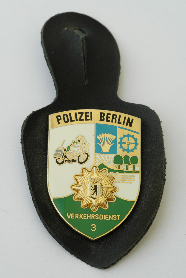 Ein schildförmiges Polizeibadge mit der Aufschrift 'Polizei Berlin' auf einem blauen Hintergrund, einem weißen Rand, einem weißen zentralen Stern und schwarzer fetter Schrift, das auf einer Oberfläche ruht.