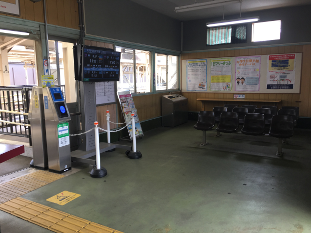 Ein belebter Bahnhofseingang mit Sitzgelegenheiten, einer Informations counter, Ticketautomaten, Deckenleuchten, Geländern und informativen Tafeln an den Wänden.