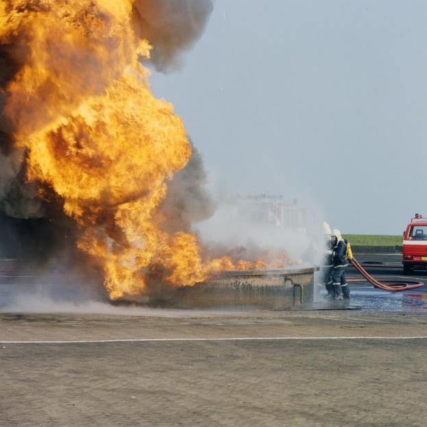 Feuerwehrfahrzeug in Flammen an der Seite einer Straße mit zwei Helmträgern, die Rohre halten, einem Fahrzeug im Hintergrund und dem Himmel.