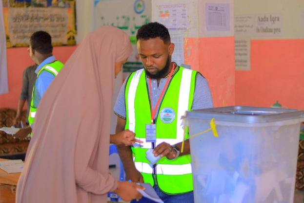 Ein Mann in einer grünen Weste und eine Frau in einem rosafarbenen Kleid stehen an einem Abstimmungstisch mit Papieren, hinter dem eine Wand mit angeklebten Papieren und ein Müllcontainer rechts zu sehen ist.