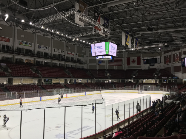 Ein Hockey-Spiel in einer großen Arena mit einem Netz in der Mitte, Zuschauern auf beiden Seiten und Banner und Fahnen an den Wänden unter einer Decke mit Lampen und Trägern, identifiziert als die St. Louis Blues Arena.