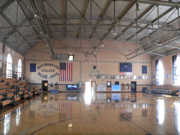 Großer Turnsaal mit Holzboden, Basketballfeld, Bänken links, Fenstern rechts, Deckenbeleuchtung und einer flaggengeschmückten Wand im Hintergrund.