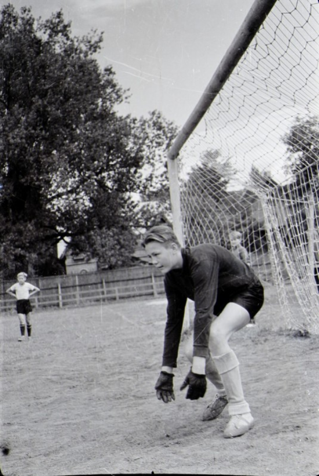 Ein Schwarz-Weiß-Foto eines jungen Jungen, der auf einem Feld Fußball spielt, mit einem Zaun, Bäumen und einem klaren Himmel im Hintergrund, anderen Kindern im Hintergrund und einer Personenschuhwerk im Vordergrund.