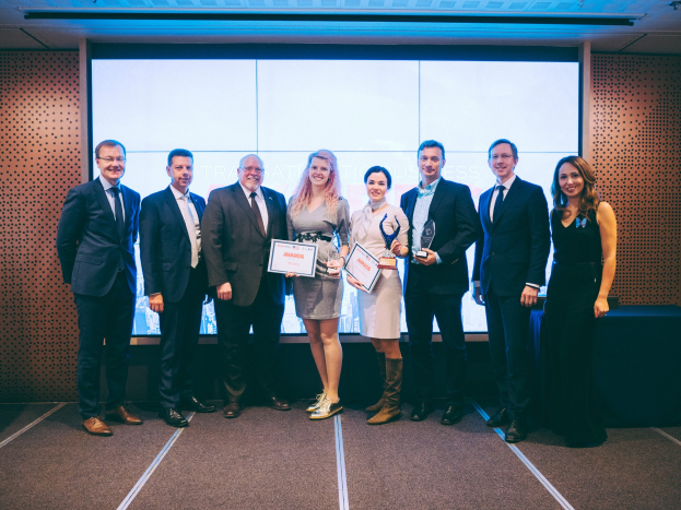 Gruppe von strahlenden Menschen mit Urkunden, die vor einem Bildschirm mit einem Tisch und einem Wandobjekt im Hintergrund stehen, Gewinner der International Business Awards 2018.