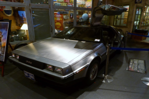 Ein DeLorean DMC-12 aus der Zurück in die Zukunft-Reihe in einem Museum ausgestellt, mit offenen Türen, umgeben von Informationsanzeigen, Bändern und verschiedenen Gegenständen.