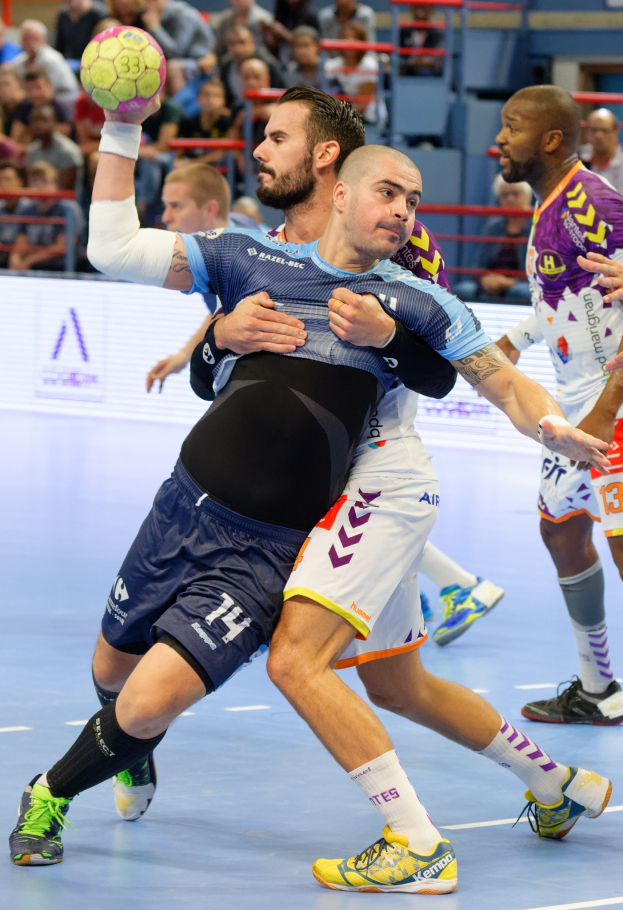 Männer beim Handballspielen in einem Stadion, einer hält den Ball, während die anderen in Bewegung sind, mit Zuschauern auf Stühlen, Geländern und einer Wand im Hintergrund.