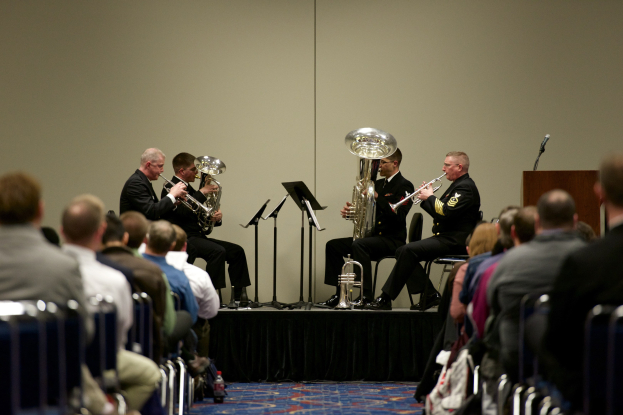 Eine Gruppe von Männern, die auf einer Bühne Blasinstrumente spielen, mit Notenständern, einem Rednerpult und einem Mikrofon, vor einem Publikum, das auf Stühlen sitzt.