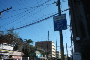 Stadtstraße mit fahrenden Autos, Strommasten mit Drähten, Gebäuden, Bäumen und Namensschildern, mit einem "Fahrer müssen den Sicherheitsgurt tragen"-Schild an einem Strommasten vor einem sichtbaren Himmel.