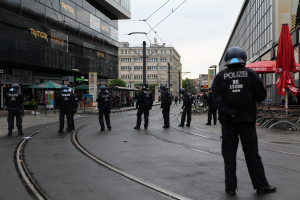 Polizeibeamte in schwarzer Uniform und Helm stehen entlang einer Straße mit Glasfenstern, Bäumen, Polen, Drähten und einem klaren Himmel, mit verstreuten Schirmen, Stühlen und Tischen.