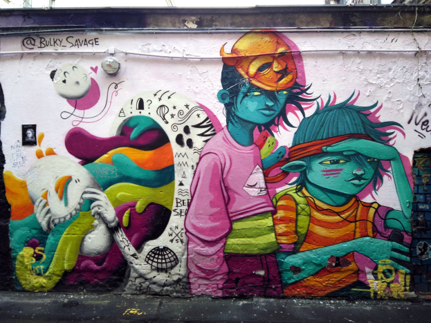 Mural an einer Gebäudeseite mit Graffiti, darunter eine Straße, Strommasten im Hintergrund und einen klaren blauen Himmel.