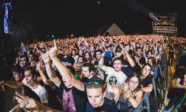 Menschenmenge auf einem Musikfestival mit erhobenen Händen, sitzend und stehend, Metallzaun auf der rechten Seite, Gebäude und Lichter im Hintergrund unter einem dunklen Himmel, mit einem Mann und einer Frau im Vordergrund.