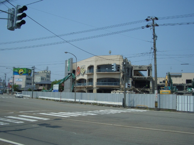 Stadtstraße mit fahrenden Fahrzeugen, Strom- und Straßenlaternen, einer Ampel auf der linken Seite, Gebäuden und Bäumen im Hintergrund und einem teilweise abgebrochenen Gebäude im Vordergrund.