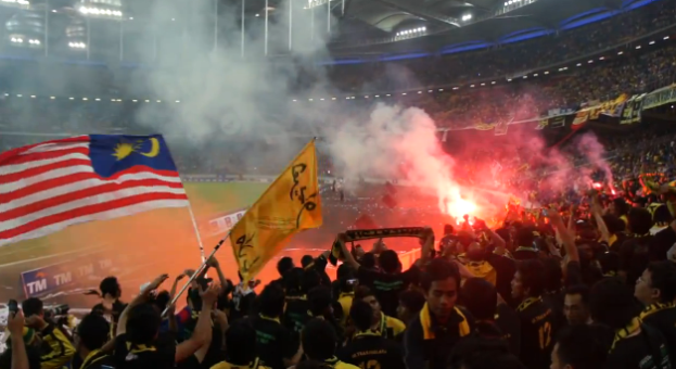 Eine große Menschenmenge in einem Stadion mit Fackeln und Rauch, einige halten Fahnen und eine Kamera, mit Schildern, die 'Malaysia vs Malaysia Live Streaming' anzeigen.