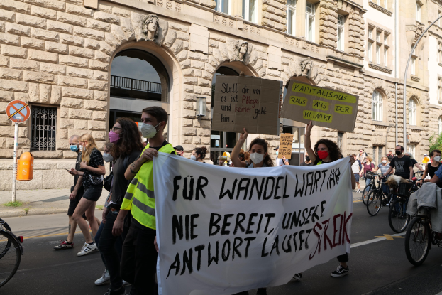 Eine Gruppe von Menschen marschiert auf der Straße in Berlin, hält Schilder und Banner und fährt mit Fahrrädern, vor einem Gebäude mit Fenstern, Bögen, Säulen, Skulpturen, Bäumen und einem Laternenmast, an einer Anti-Kriegs-Demonstration teilnehmend.