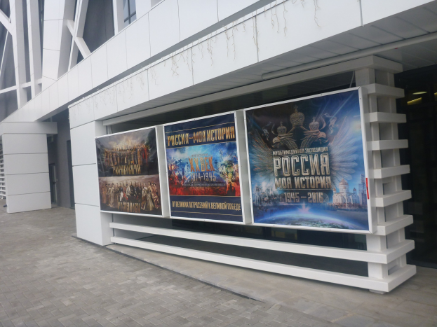 Großes Werbeplakat an einem Gebäude für die Eröffnungszeremonie der Olympischen Spiele in Moskau, mit sichtbarem Boden und rechter Beleuchtung.