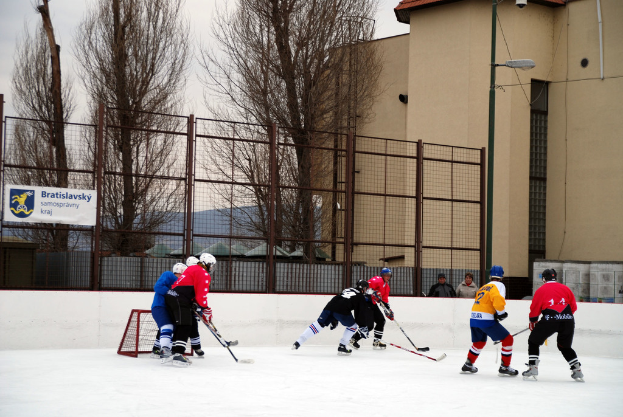 Menschen beim Eisschnellaufen auf einer Eisbahn mit Gebäuden, Bäumen, einer Straßenlaterne, einem Namensschild und Zäunen im Hintergrund bei klarem Himmel.