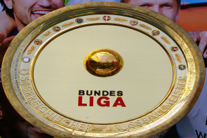 Ein Mann hält einen goldenen Schild mit der Aufschrift "Bundesliga" hoch, im Hintergrund ein Banner mit drei Personen.