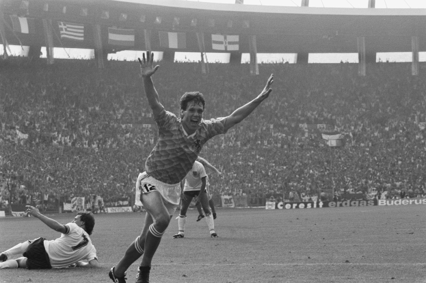 Schwarzes und weißes Foto eines Fußballspielers, der nach einem Tor im Stadion feiert, umgeben von Teamkollegen und Stab, mit Stadiontafeln, Fahnen und Pfosten im Hintergrund.
