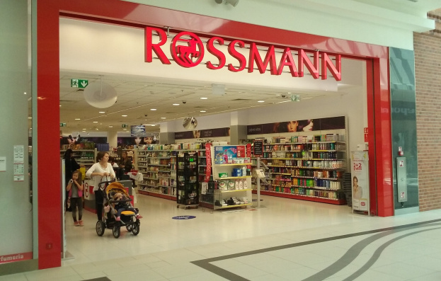Ein Rossmann-Laden in einem Einkaufszentrum mit Deckenleuchten, Schildern, Menschen und Regalen voller verschiedener Gegenstände, eine Person mit einem Kinderwagen.