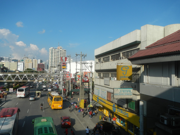 Eine belebte Stadtstraße mit Autos, Bussen, Fußgängern auf dem Gehweg, Strommasten, Gebäuden mit Schildern, einer Brücke, Bäumen und einem bewölkten Himmel.