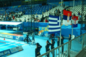 Eine Gruppe von Menschen in Uniformen auf einem Swimmingpool stehend, umgeben von Metallzaun, Flaggen, Kamerasetup, Möbeln, einem Banner und einem überdachten Gebäude mit Deckenbeleuchtung.