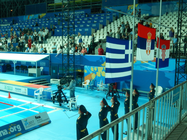Eine Gruppe von Menschen in Uniformen auf einem Swimmingpool stehend, umgeben von Metallzaun, Flaggen, Kamerasetup, Möbeln, einem Banner und einem überdachten Gebäude mit Deckenbeleuchtung.