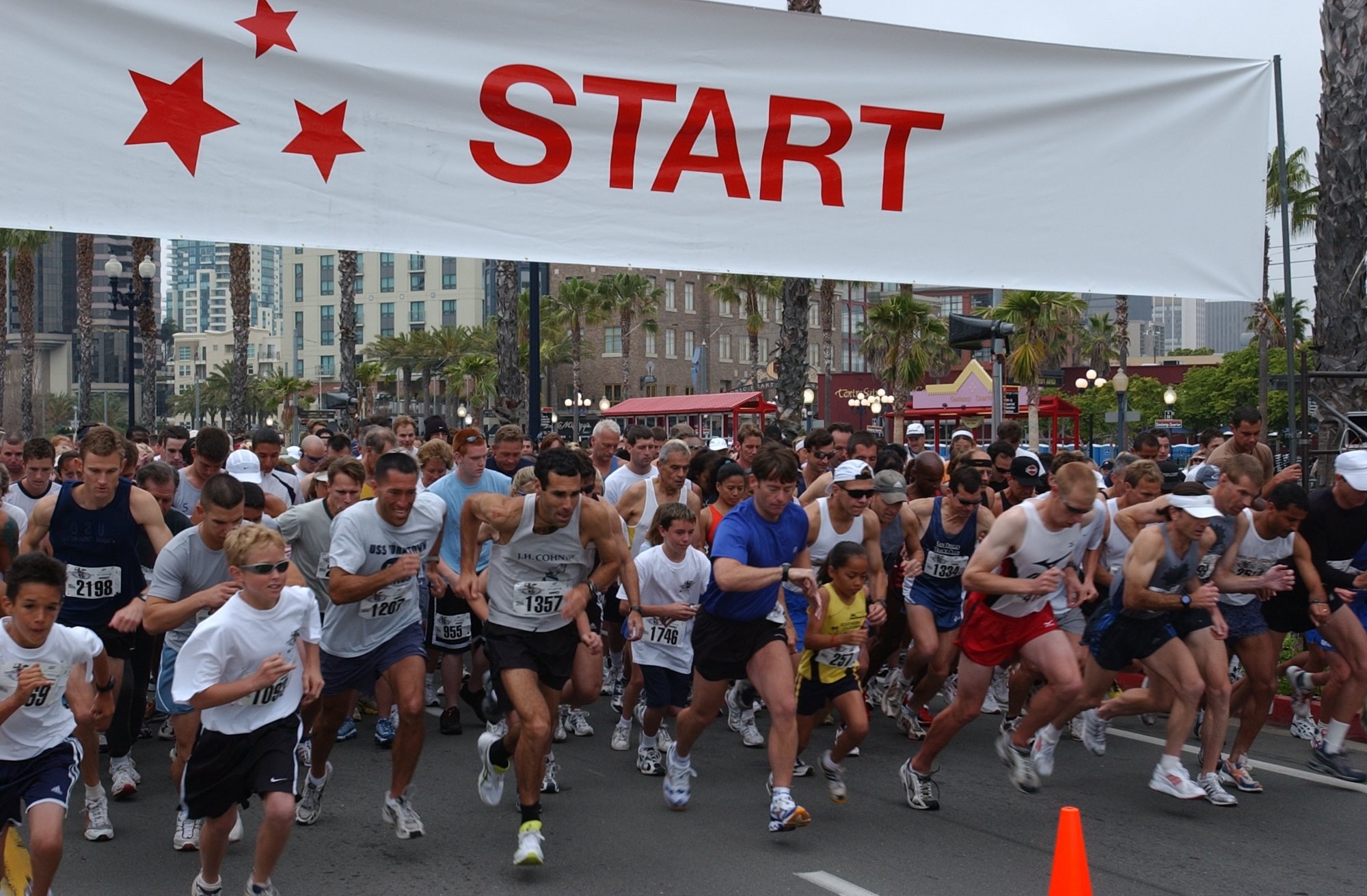 Gruppe von Läufern bei einem Marathon mit einem Verkehrskegel im Vordergrund und einem Banner im Hintergrund, umgeben von Bäumen, Laternenmasten, Gebäuden und einem klaren blauen Himmel.