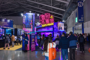 Eine große Gruppe von Menschen steht vor einem Stand auf einer Messe, einige halten Taschen, rechts ist eine Wand zu sehen. Der Stand präsentiert Schilder mit der Aufschrift "Indie Game Development Award 2019", Bildschirme, Lampen und weitere Gegenstände im Hintergrund.