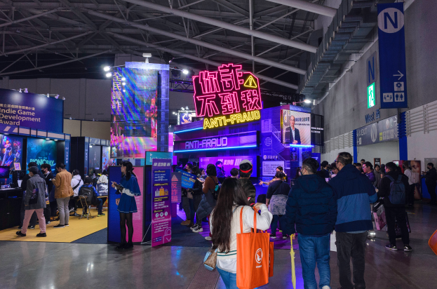 Eine große Gruppe von Menschen steht vor einem Stand auf einer Messe, einige halten Taschen, rechts ist eine Wand zu sehen. Der Stand präsentiert Schilder mit der Aufschrift "Indie Game Development Award 2019", Bildschirme, Lampen und weitere Gegenstände im Hintergrund.
