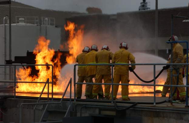 Eine Gruppe von Feuerwehrleuten in Helmen steht auf dem Dach eines Gebäudes, hält Rohre, mit Geländern, Treppen, einem anderen Gebäude und dem Himmel im Hintergrund.
