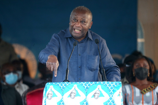 Ein Mann in einem blauen Hemd spricht in zwei Mikrofone an einem Rednerpult, mit sitzenden Zuschauern und einer Wand im Hintergrund.