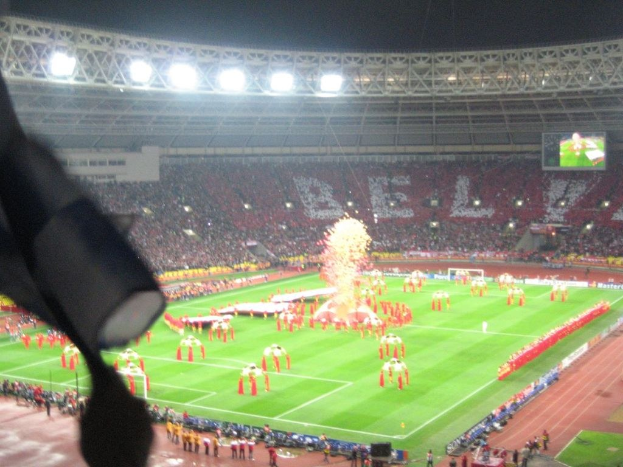 Ein Stadion mit Flutlicht, Eisengittern, Zuschauern, stehenden Personen, Werbetafeln, Anzeigetafeln, Überwachungskamera, Fahnen und Deko-Elementen.
