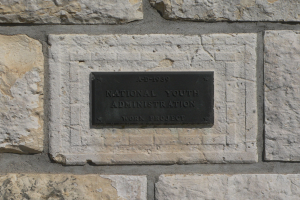 Steinwand mit einem rechteckigen Schild, auf dem in weißer, fetter, zentrierter Schrift 'National Youth Administration' steht.