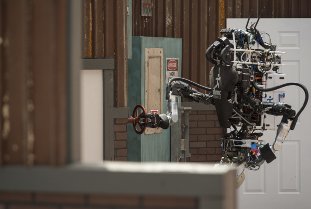 Humanoider Roboter mit ausgestreckten Armen und geneigtem Kopf vor einem Gebäude mit einer Tür und einer Wand im Hintergrund.